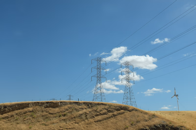 A photograph of a landscape with electric towers and a wind turbine on a hill under a clear blue sky.