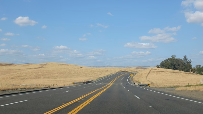 A paved highway curves through dry, golden grasslands under a bright blue sky with scattered white clouds.