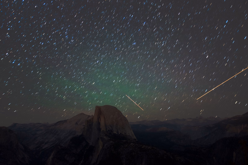A long-exposure photograph captures a meteor shower streaking across a dark night sky above a silhouetted mountain range.