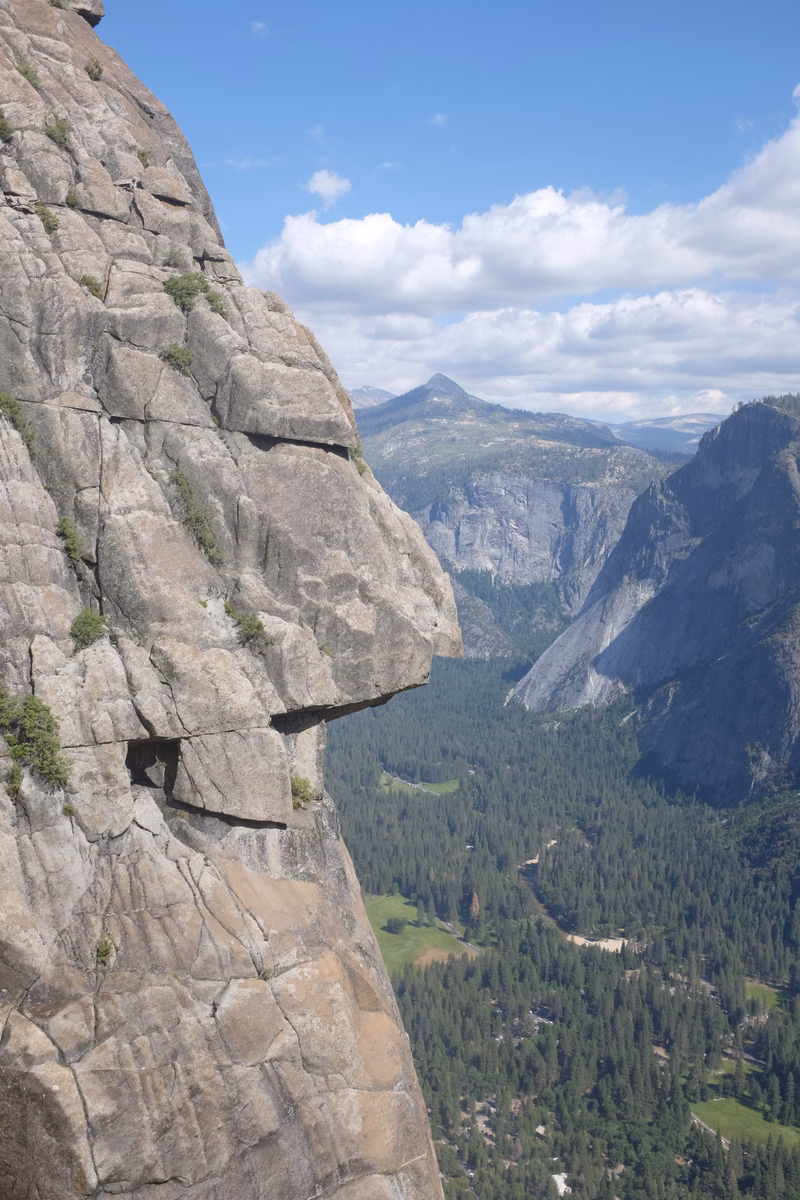 A massive gray rock formation overlooks a dense evergreen forest nestled between steep mountain cliffs under a partly cloudy blue sky.