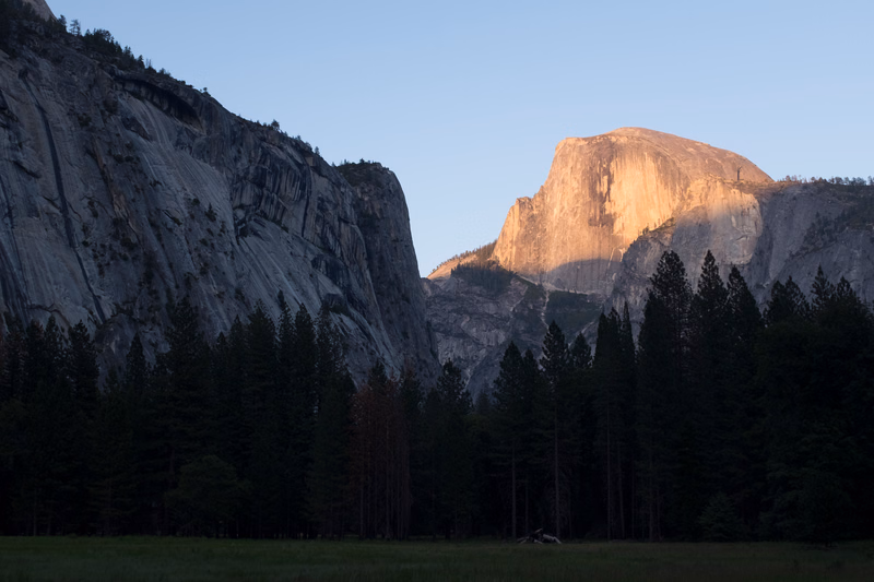 Half Dome mountain is illuminated by the setting sun behind a dark forest and rocky cliff face.