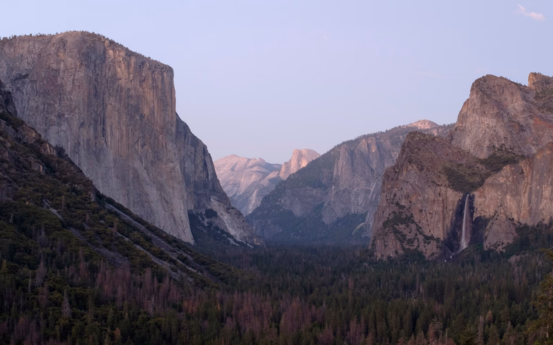 Towering granite cliffs frame a deep valley filled with dense evergreen forest, with a waterfall visible on the right.