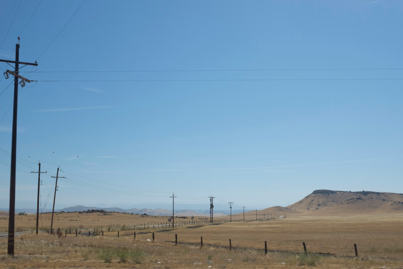 A wide, open landscape under a clear blue sky, featuring several utility poles and power lines stretching across a dry field.