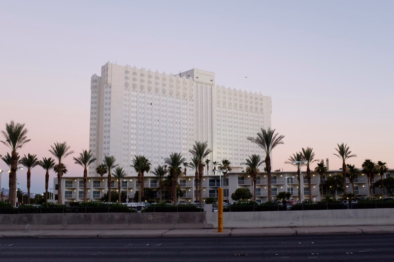 A large white hotel building with many windows is seen behind a row of palm trees near the coast during twilight.
