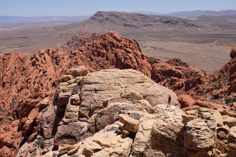 A hiker stands on a large, layered rock formation overlooking a vast desert landscape with distant mountains under a clear sky.