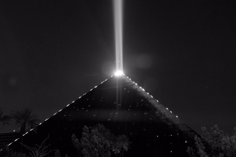 A bright laser beam shoots vertically from the apex of a large pyramid structure against a dark night sky.