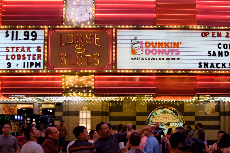 A brightly lit commercial building features signs advertising "$11.99 STEAK & LOBSTER 9 - 11PM," "LOOSE $ SLOTS," and "DUNKIN' DONUTS AMERICA RUNS ON DUNKIN'".