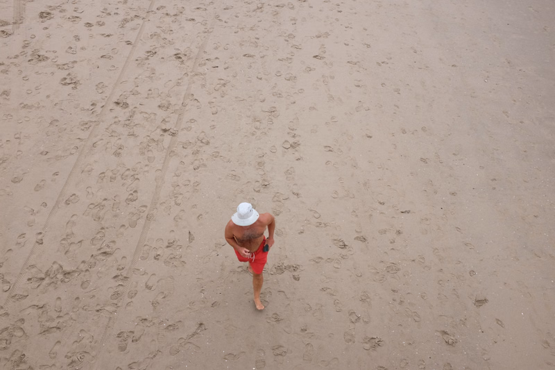 A man wearing a white hat and red shorts walks across the sandy beach.