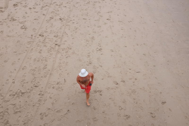 A man in a white hat and red shorts walking on a sandy beach.