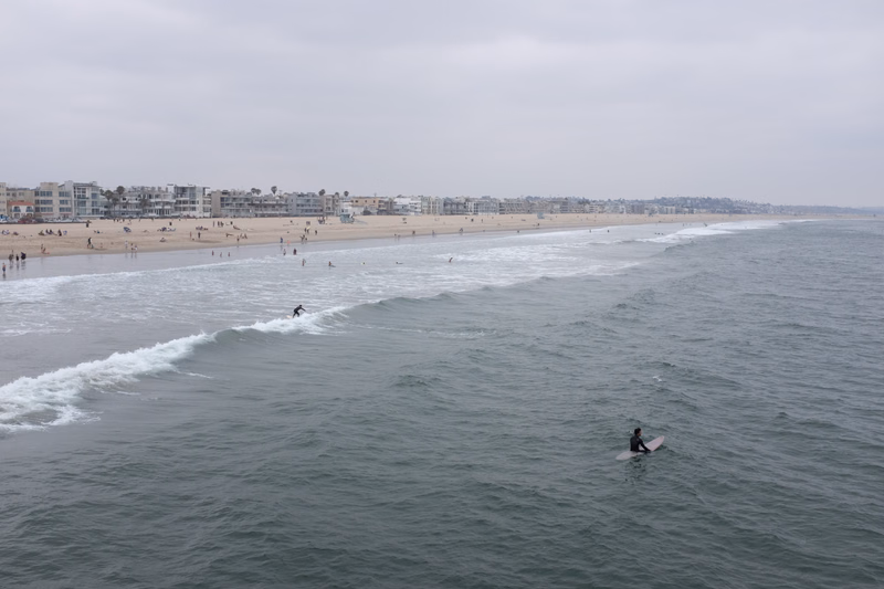 A surfer rides a wave near the shore of a long sandy beach with buildings lining the coast under an overcast sky.