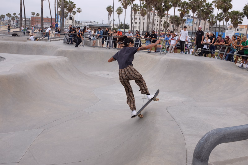 A skateboarder in patterned pants performs a trick in a large concrete skatepark with spectators watching from the background.