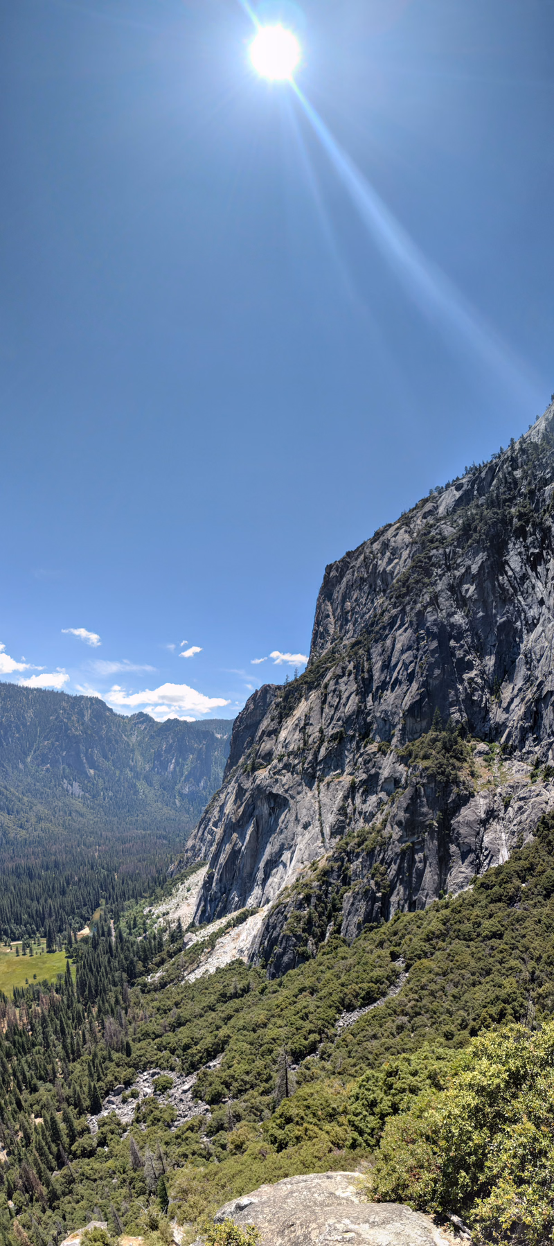 A bright sun shines over a steep rocky mountain face covered in green forest under a clear blue sky.