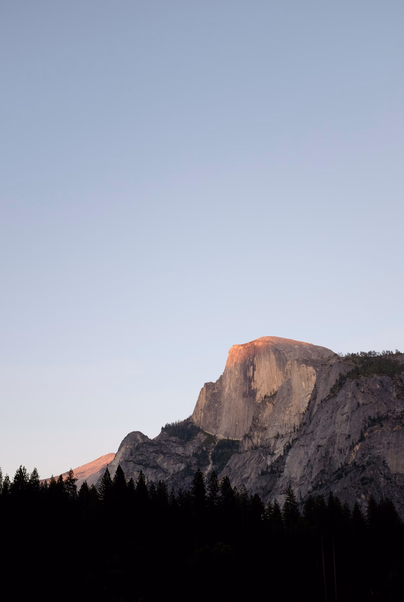 A large granite mountain peak rises above a dark evergreen forest under a pale sky.